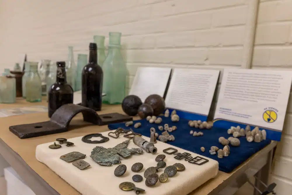 Display of historical artifacts on a table with glass bottles, metal tools, and coins in a museum exhibit setting.