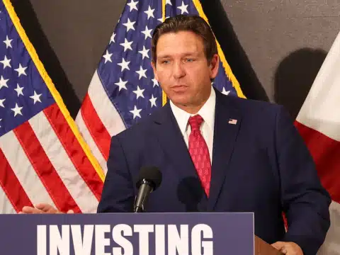 Male politician in a dark blue suit and red tie speaks at a podium with a microphone, backdrop of American flags and a banner reading 'INVESTING' behind him.