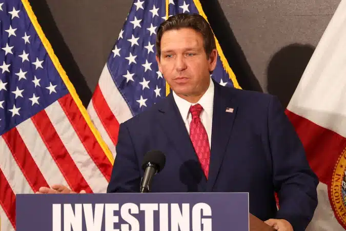 Male politician in a dark blue suit and red tie speaks at a podium with a microphone, backdrop of American flags and a banner reading 'INVESTING' behind him.