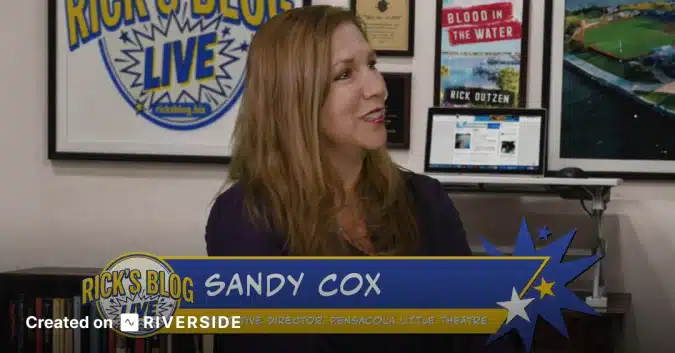 Sandy Cox seated at a desk in a studio, with a blue banner reading 'Sandy Cox' in the foreground and posters on the wall behind her.