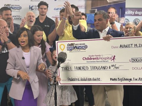 Group of people celebrate as a large ceremonial check is presented for $300,000 to Childrens Hospital, with a microphone in front and clapping in the background.