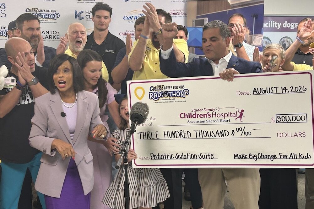 Group of people celebrate as a large ceremonial check is presented for $300,000 to Childrens Hospital, with a microphone in front and clapping in the background.